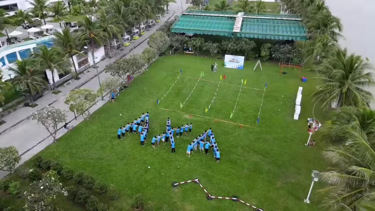 Team-building Trip 2023 31 Aerial view of a lush green lawn with people in blue shirts forming the letters APA. This meticulously organized area, possibly planned with civil engineering precision, is sectioned off with markers leading to a net for a team event. Palm trees and a body of water gracefully envelop the scene.